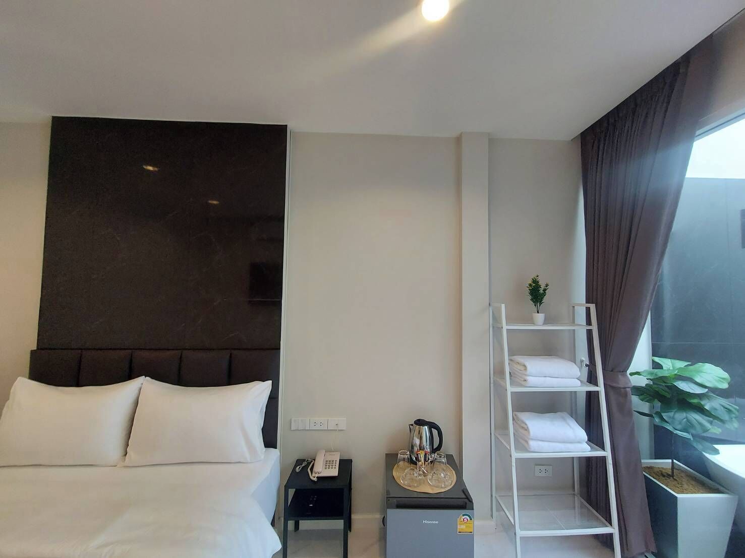 Een moderne hotelkamer met een groot bed, een nachtkastje met een telefoon en een waterkoker, een klein plankje met handdoeken, en een raam met gordijnen.
