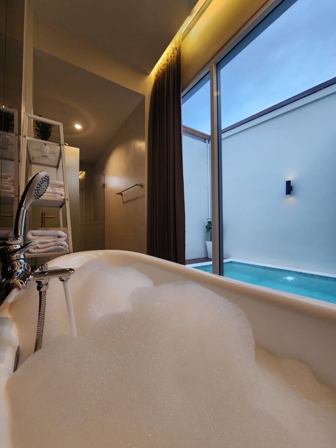 A modern bathroom with a large bathtub filled with bubbles, a shower area, and a view of a swimming pool through a large window.
