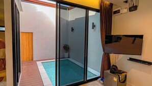 A modern room with a sliding glass door leading to a balcony with a pool, a flat-screen TV mounted on the wall, and a small trash bin. - KVILLA (Bangkok)