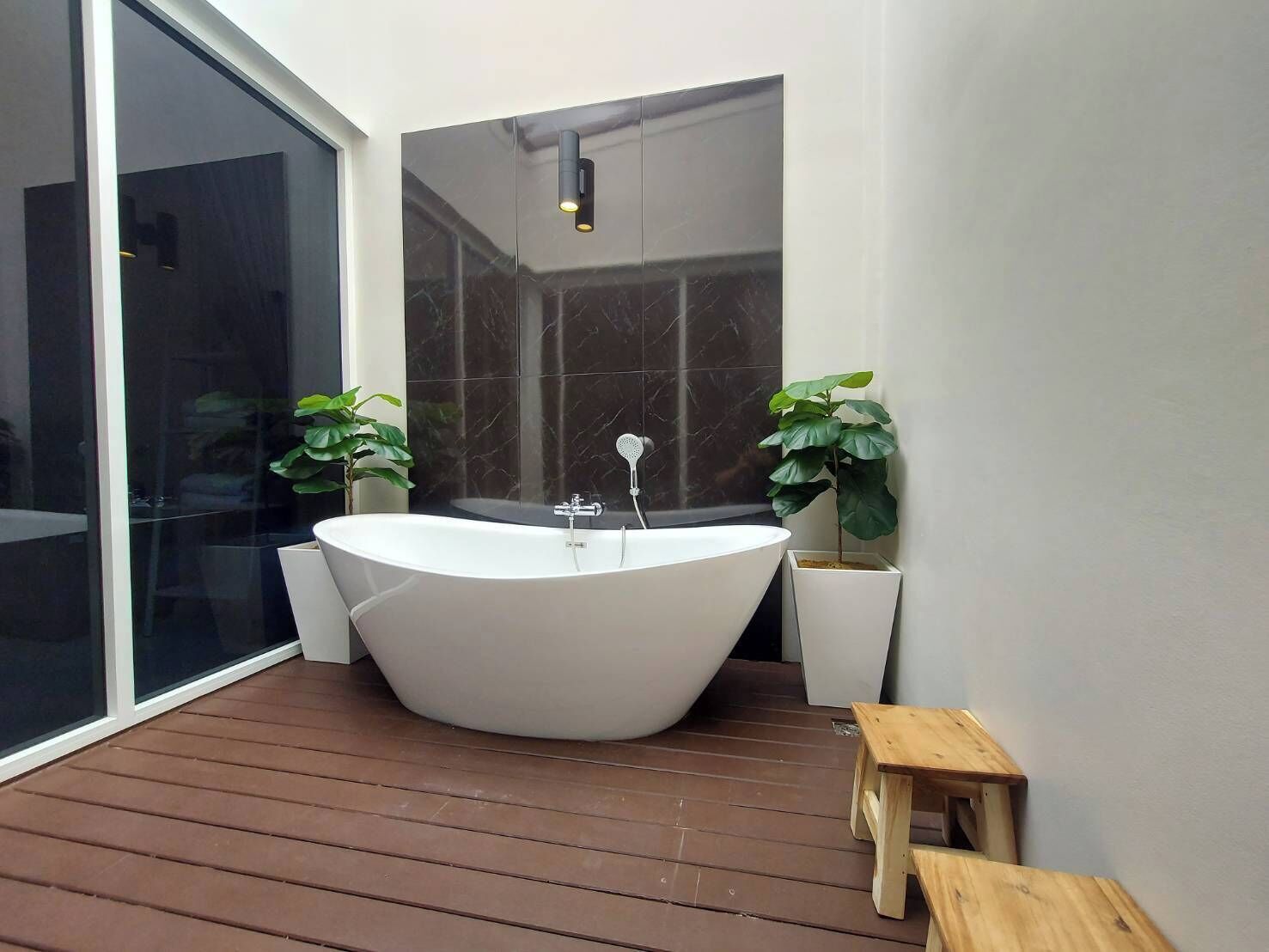 A modern bathroom with a freestanding bathtub, a glass door leading to another room, and two potted plants.