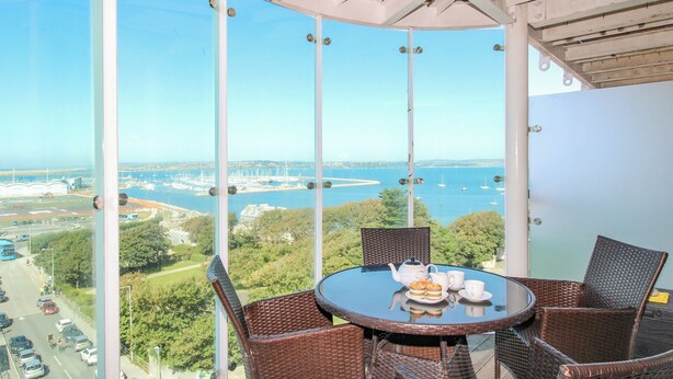 A balcony with wicker chairs and a table, overlooking a marina and a bridge.