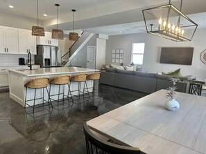 A modern kitchen with a central island, bar stools, a dining table, and a staircase leading to a living area.