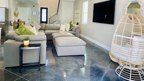 Un salon moderne avec un canapé d’angle, une petite table, un grand téléviseur à écran plat et un fauteuil suspendu décoratif.
