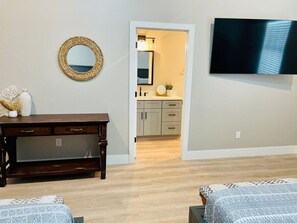 A room with a wooden desk, a television mounted on the wall, and a doorway leading to a kitchen area.