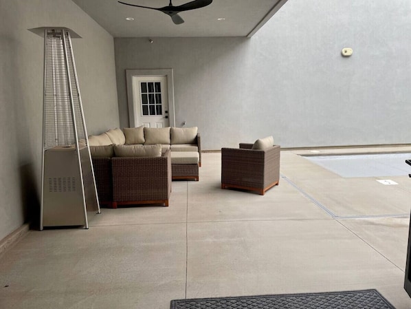 A modern living room with a sofa, two wicker chairs, a standing heater, and a pool area.
