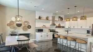 Une cuisine moderne avec des meubles blancs, un îlot central doté de tabourets de bar, une table à manger avec des chaises et des luminaires suspendus.