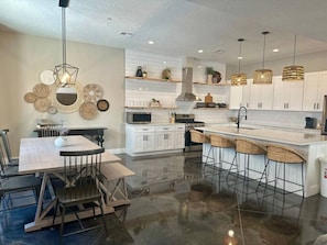 A modern kitchen with white cabinets, a central island with bar stools, a dining table with chairs, and pendant lighting.