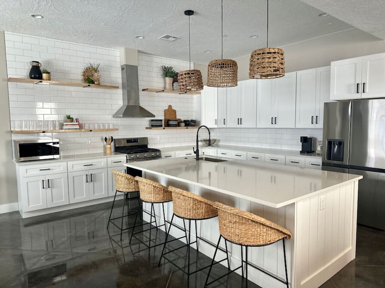 A modern kitchen with white cabinetry, a large island with bar stools, a stainless steel refrigerator, and hanging wicker light fixtures.