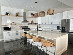 A modern kitchen with white cabinetry, a large island with bar stools, a stainless steel refrigerator, and hanging wicker light fixtures.