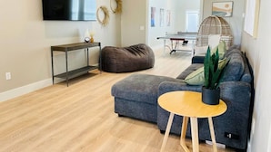 Un salon moderne avec un canapé gris, une petite table en bois, un fauteuil poire, un téléviseur et une table de ping-pong.