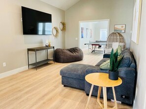 A modern living room with a grey sofa, a small wooden table, a bean bag, a TV, and a ping pong table.
