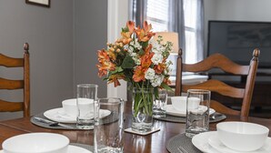Ein Esstisch ist gedeckt mit weißen Tellern, Schüsseln und Gläsern, dazu ein Blumenstrauß in einer Vase.