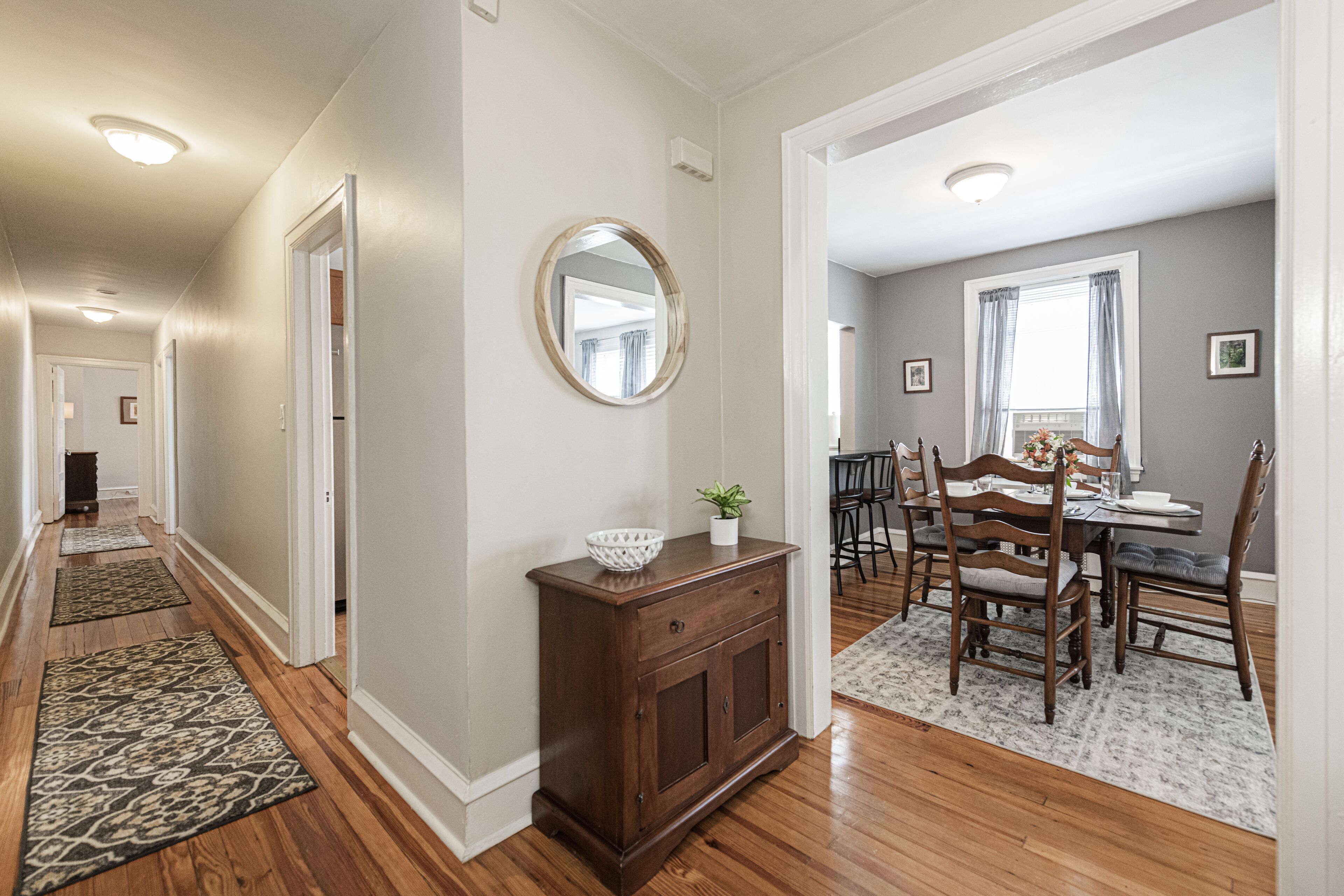 A hallway with wooden flooring, a dining area with a table and chairs, and a mirror on the wall.