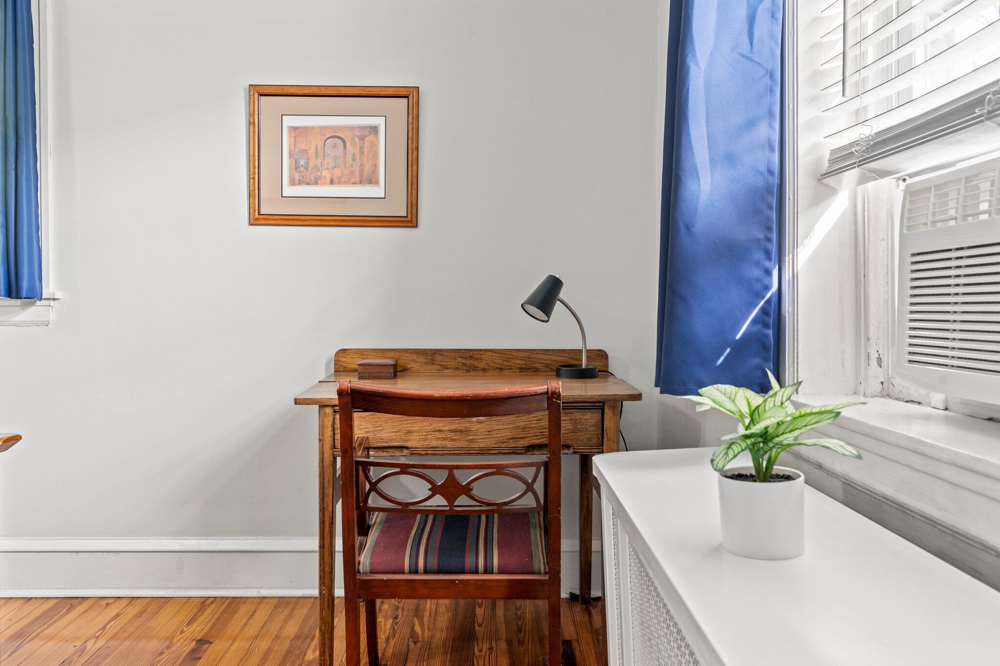 Una habitación con un escritorio de madera, una silla, un cuadro en la pared, una planta en maceta en el alféizar de la ventana y una cortina azul.