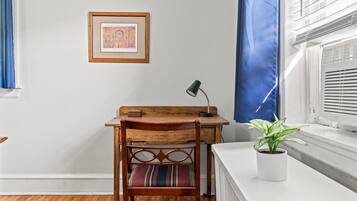 Una habitación con un escritorio de madera, una silla, un cuadro en la pared, una planta en maceta en el alféizar de la ventana y una cortina azul.