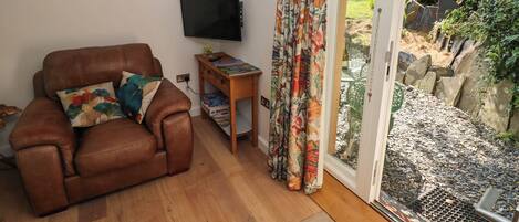 Uma sala de estar com sofá de couro, televisão de tela plana, mesa lateral de madeira e porta de vidro deslizante que dá para um jardim.