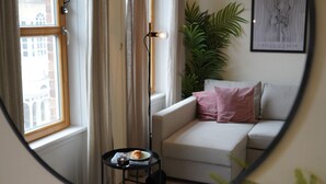 Een knusse woonkamer met een bank, een kleine tafel met een croissant en een drankje, een vloerlamp en een grote plant.