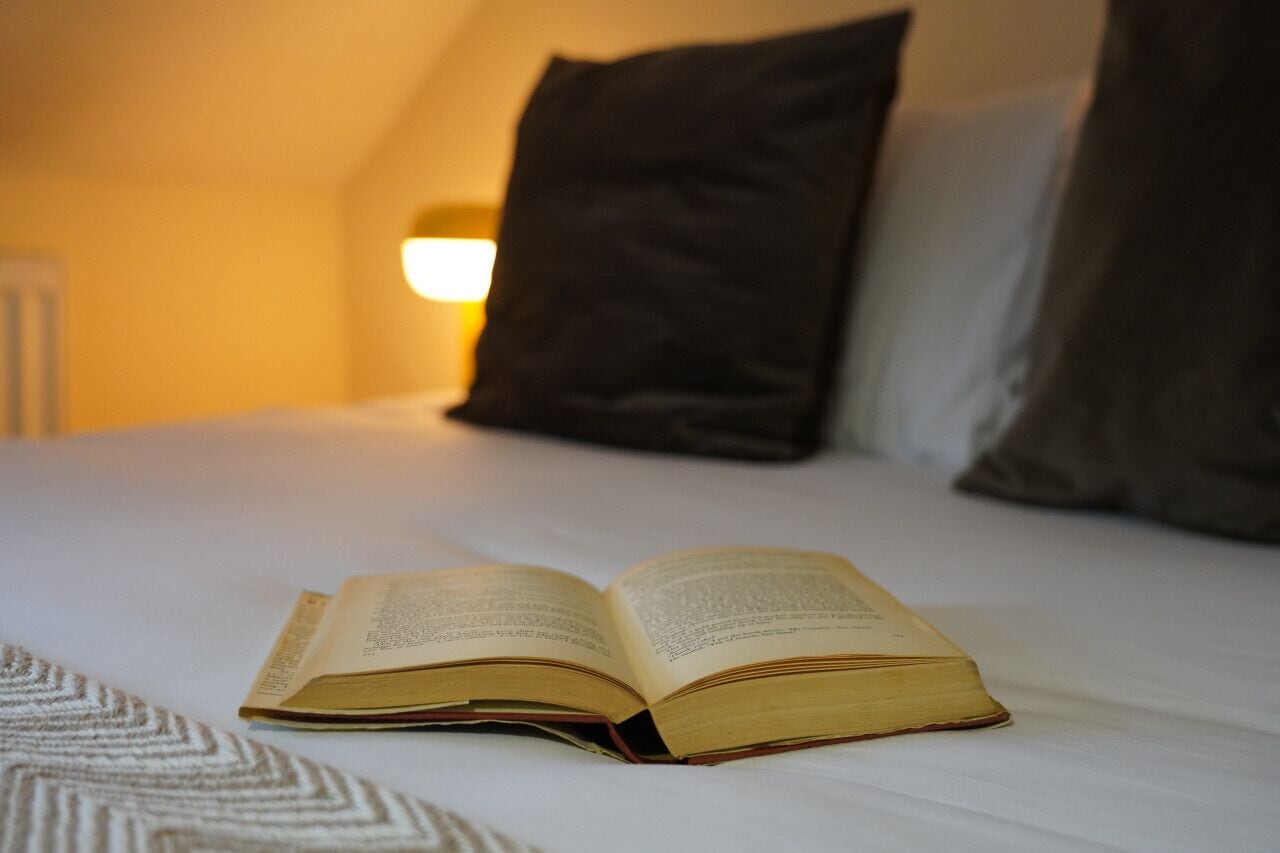 An open book on a bed in a room with a lamp and pillows.