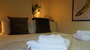 Een net opgemaakt bed met witte lakens, twee donkere kussens, een opgevouwen witte handdoek en een wandlamp.