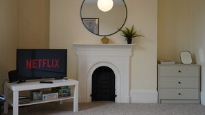 Een woonkamer met een open haard, een televisie waarop 'NETFLIX' staat, een ronde spiegel en een kleine plant op de schouw.