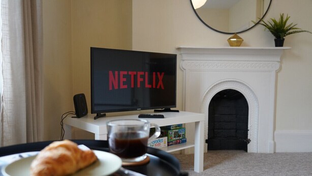 Een woonkamer met een televisie waarop het Netflix-logo te zien is, een open haard en een dienblad met een croissant en koffie.