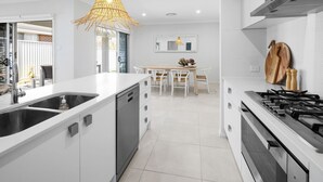 A modern kitchen with white cabinetry, a dishwasher, a stove, and a dining area with a table and chairs. - Spoondrift - 5 Mins Walk to Beach (South West Rocks)
