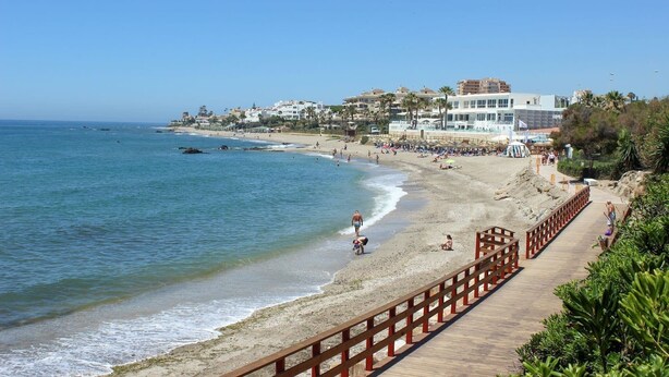A beach with a wooden walkway, clear blue water, and buildings in the background. - Modern 2 bed apartment Riviera close to the beach (Mijas Costa)