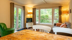 A cozy living room with a wooden table, a green chair, a white sofa, and a television on a white cabinet. Large windows with curtains allow natural light and views of greenery outside.