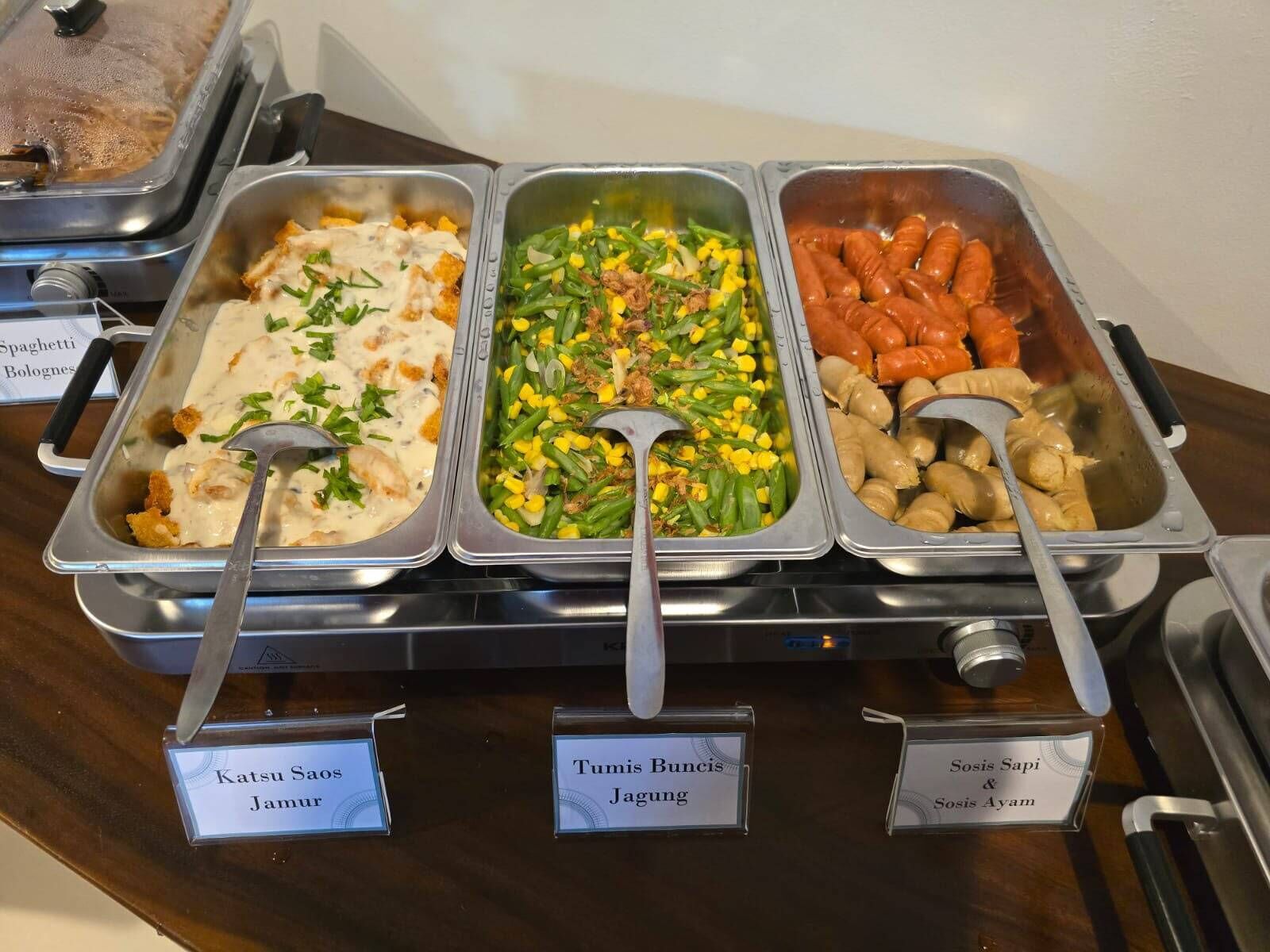 A buffet with labeled dishes including spaghetti bolognese, green beans with corn, and sausages.