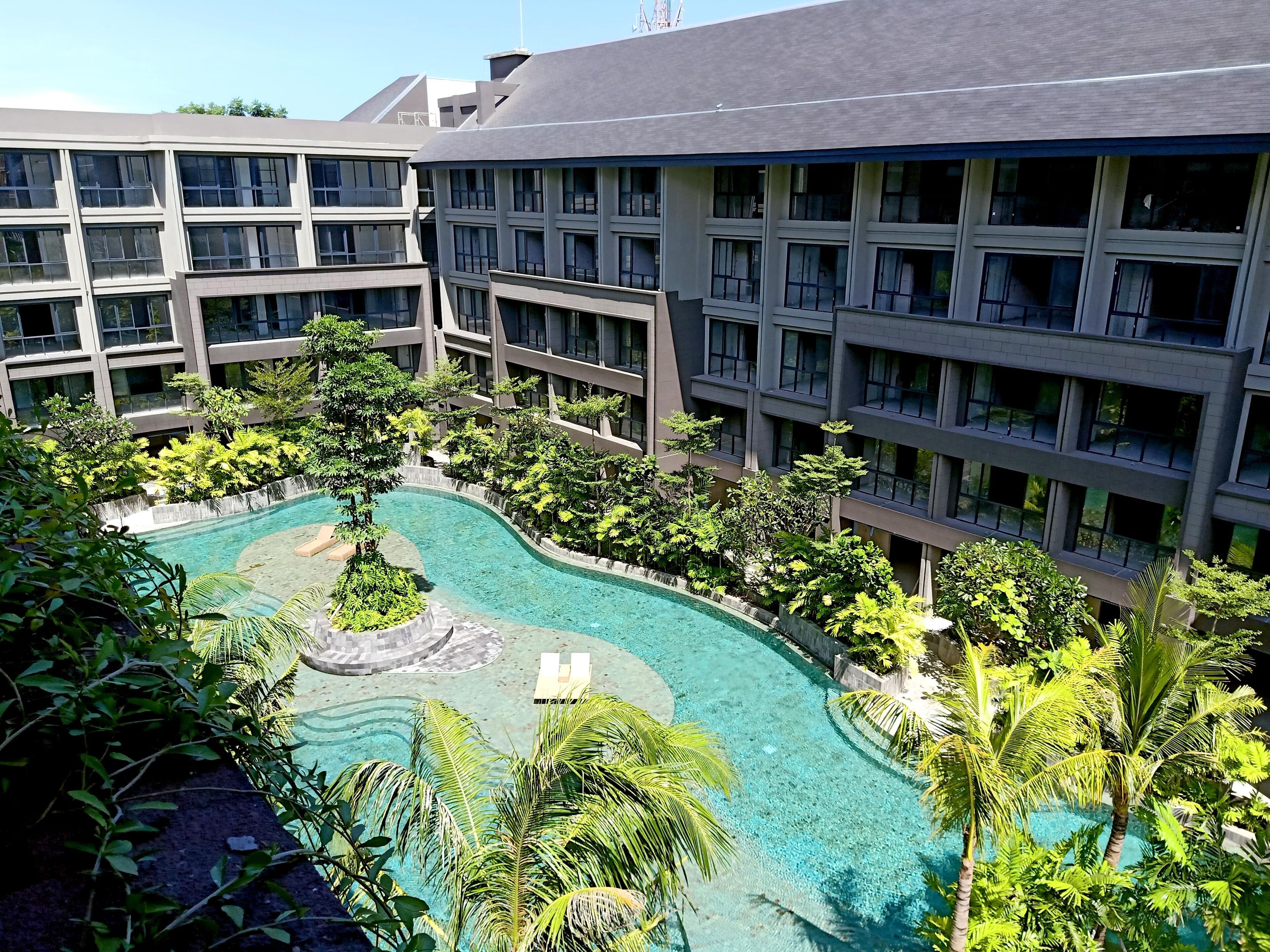 A modern hotel with a central swimming pool surrounded by lush greenery and multiple balconies.