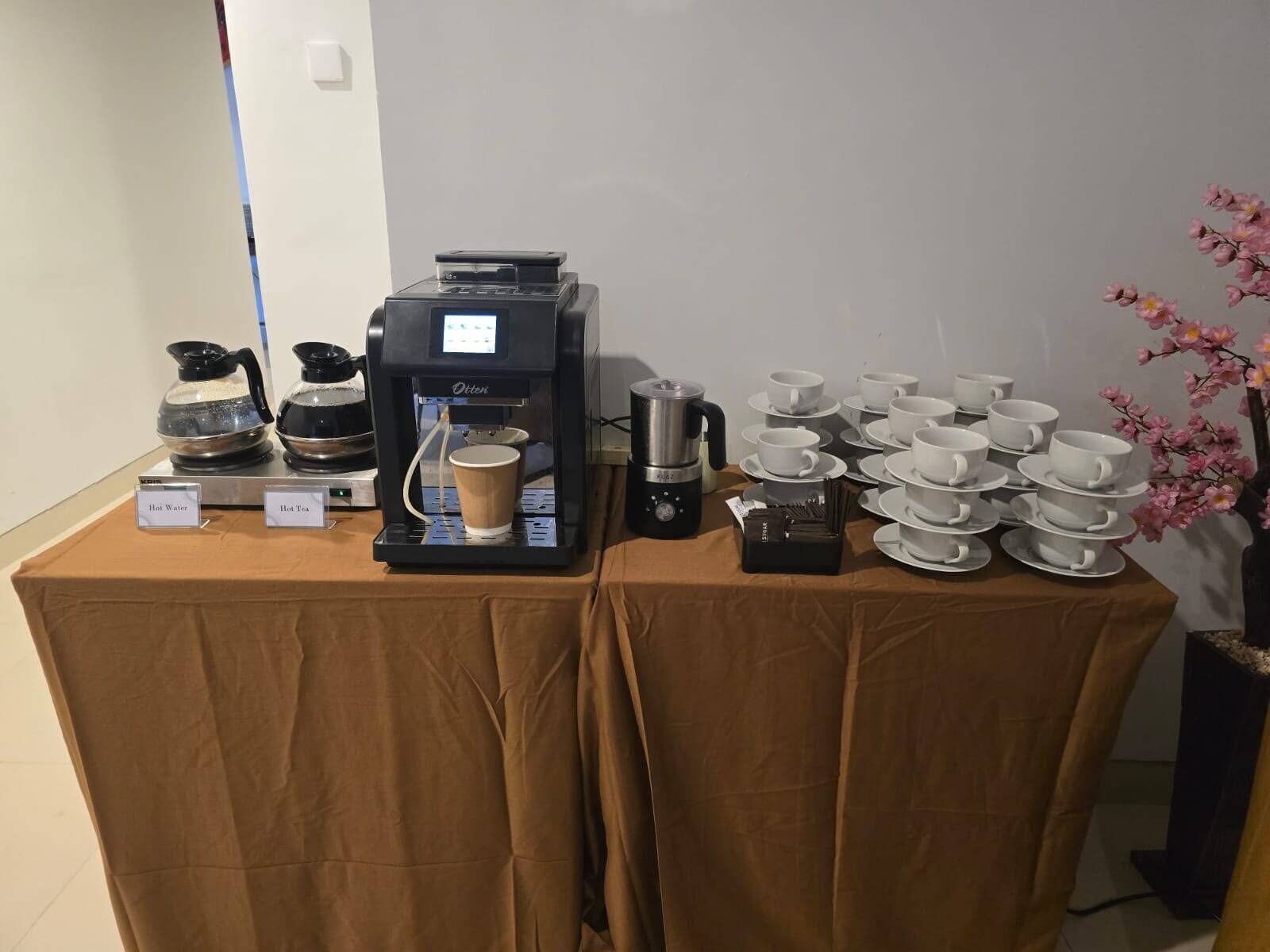 Una estación de café con una máquina de café, dispensadores de agua caliente y té, y una pila de tazas y platillos blancos.