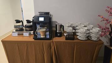 Una estación de café con una máquina de café, dispensadores de agua caliente y té, y una pila de tazas y platillos blancos.