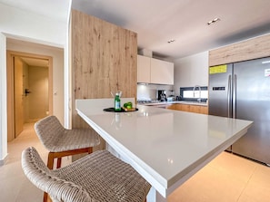 A modern kitchen with a central island, two wicker chairs, stainless steel appliances, and wooden cabinetry.