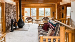 A cozy cabin interior with a stone wall, a fireplace, wooden furniture, and large windows offering a view of trees. - Stunning Views & Starry Skies Deck & BBQ (Pine)