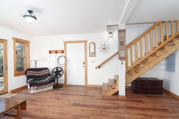 Una habitación con suelo de madera, una escalera, un ventilador, una puerta y una lámpara fijada a la pared.