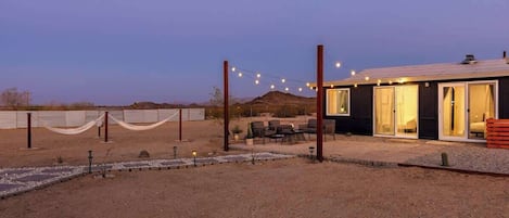 Paisaje desértico con un edificio que tiene un patio con sillas y luces colgantes.