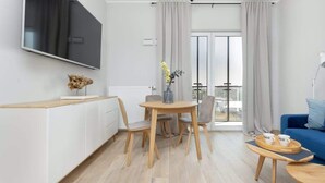 A modern living room with a TV mounted on the wall, a dining area with a table and chairs, a sofa, and a balcony with curtains. - Elegant Apartment with Balcony | Parking and City View (Warszawa)