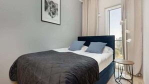 A bedroom with a bed, a small table, a window, and a framed picture on the wall. - Elegant Apartment with Balcony | Parking and City View (Warszawa)