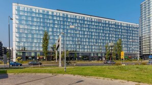 A modern building with a glass facade, a parking lot, and a road with cars. - Elegant Apartment with Balcony | Parking and City View (Warszawa)