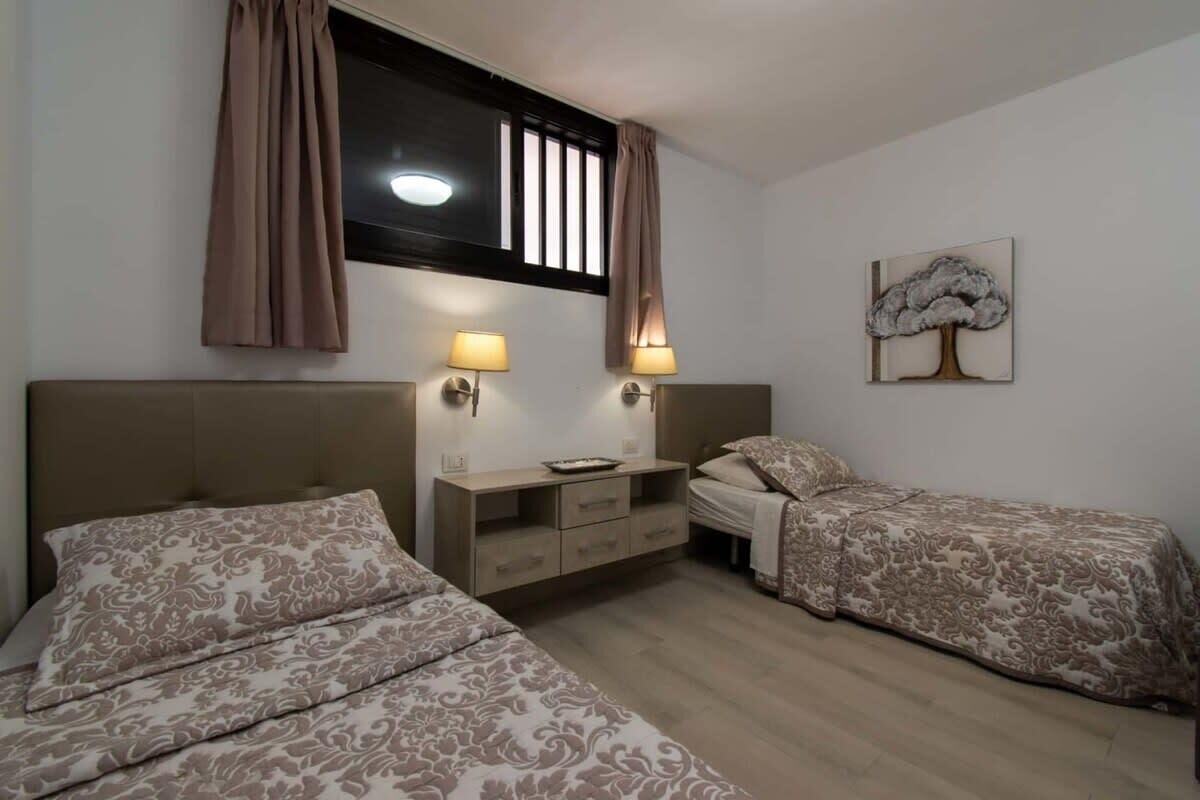 A bedroom with two beds, a bedside table, a lamp, a window with curtains, and a wall-mounted artwork.