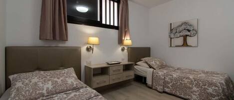 A bedroom with two beds, a bedside table, a lamp, a window with curtains, and a wall-mounted artwork.