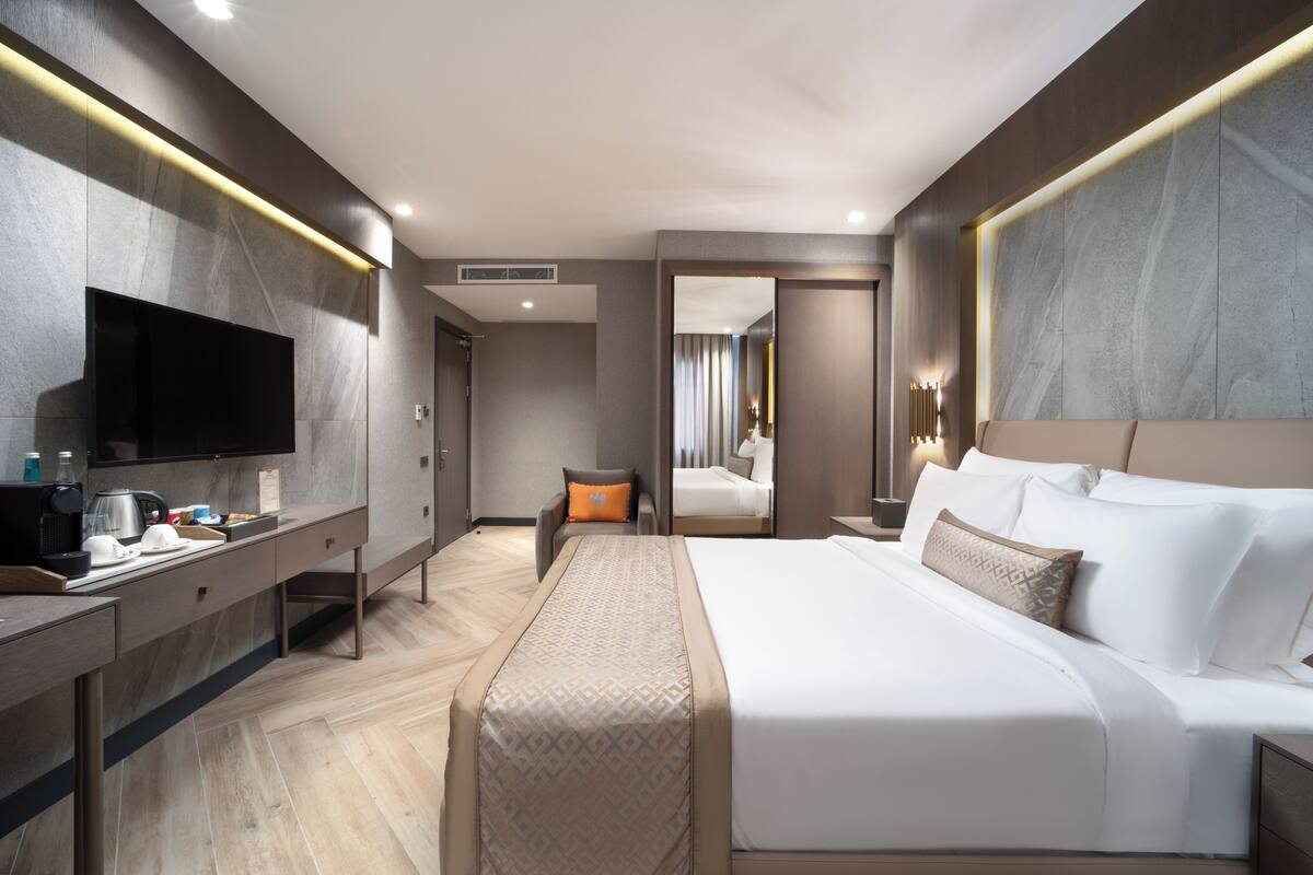 A modern hotel room with a large bed, a flat-screen TV mounted on the wall, a desk with a coffee maker, and a small seating area with a chair and a table.