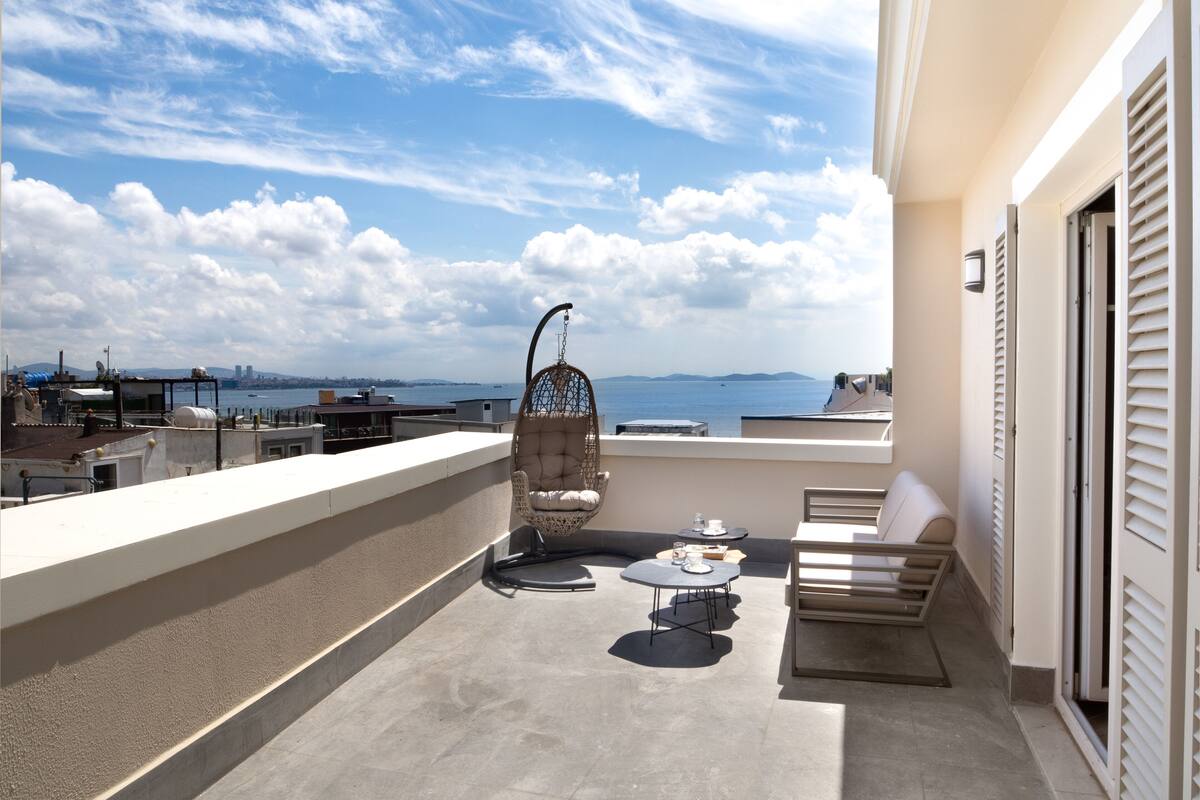 A rooftop terrace with a hanging chair, a small table with cups, and a view of the sea and distant mountains.