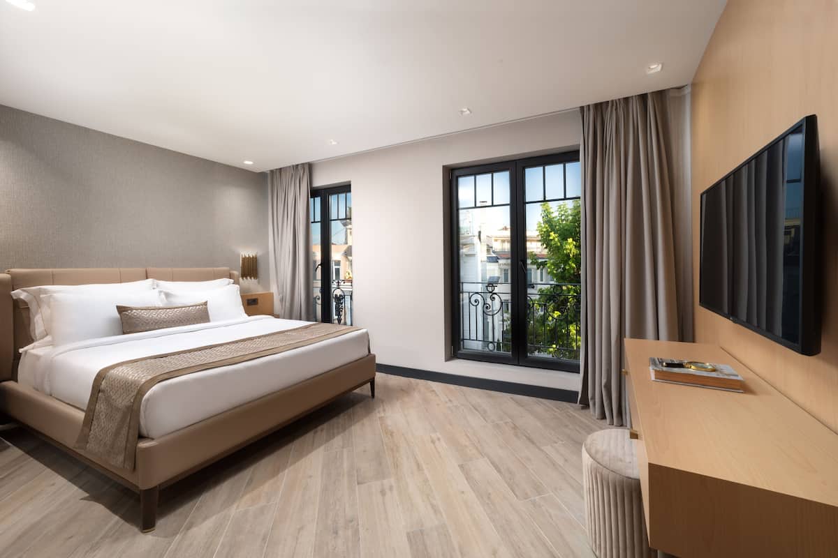 A modern hotel room with a large bed, a desk, a TV, and a balcony with curtains.