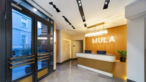 Un área de recepción con un mostrador de madera, un gran letrero iluminado que dice "MULA" y una puerta de cristal que da a un espacio exterior.