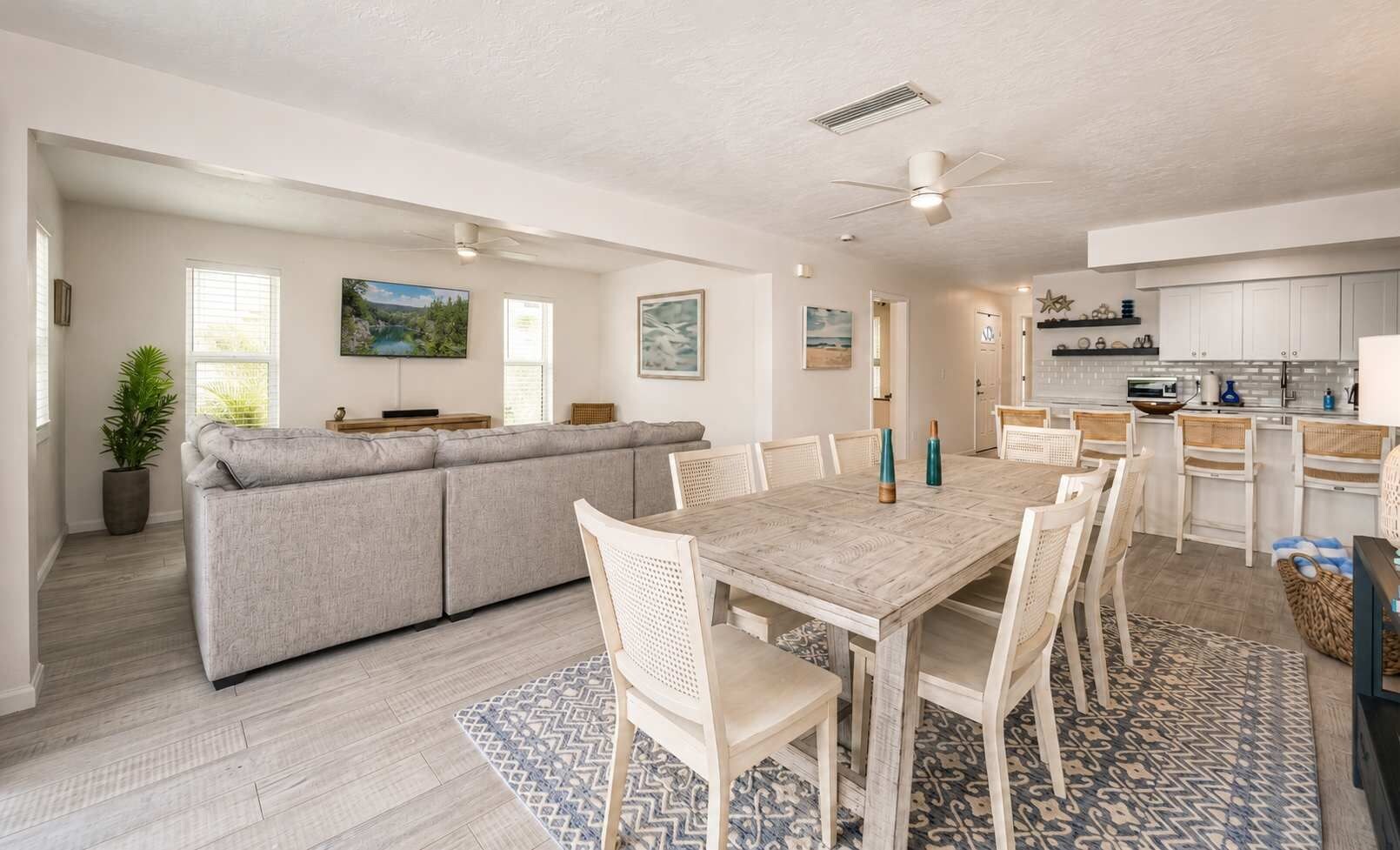 Un vaste espace de vie comprenant un canapé, une table à manger et des chaises, une cuisine avec des meubles blancs et un ventilateur de plafond.