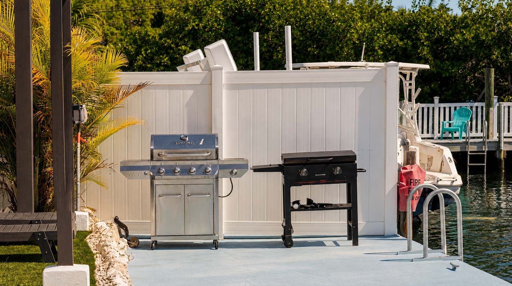 A waterfront outdoor area with a grill and a table, a dock with a boat, and a white fence.