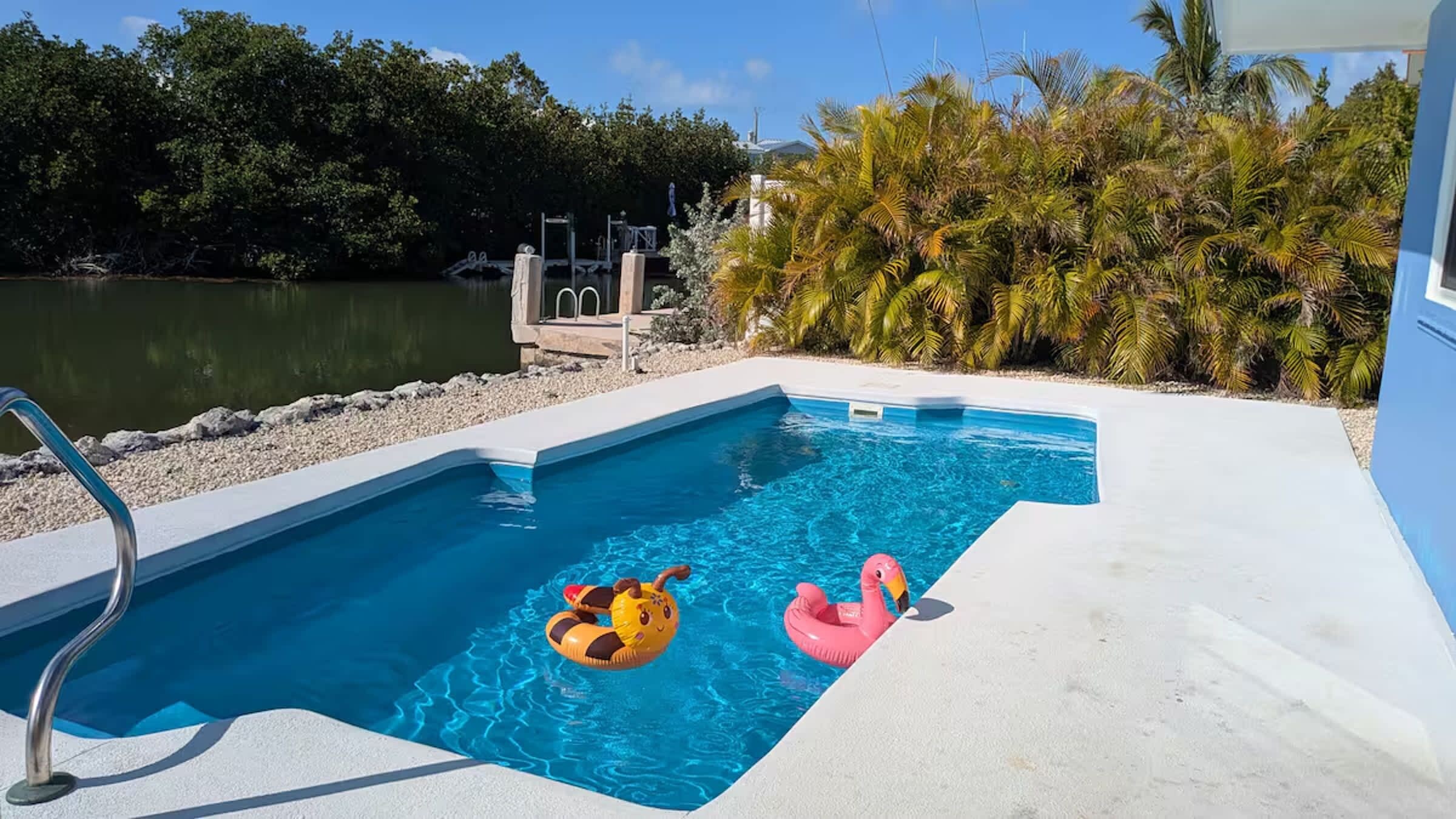 A swimming pool with two inflatable toys, surrounded by a concrete deck and palm trees.