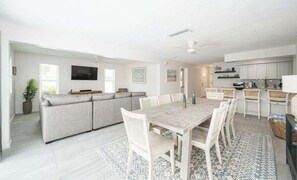 Een ruime woonkamer met een grijze bank, een witte eettafel met stoelen, een flatscreen-tv en een keuken op de achtergrond.