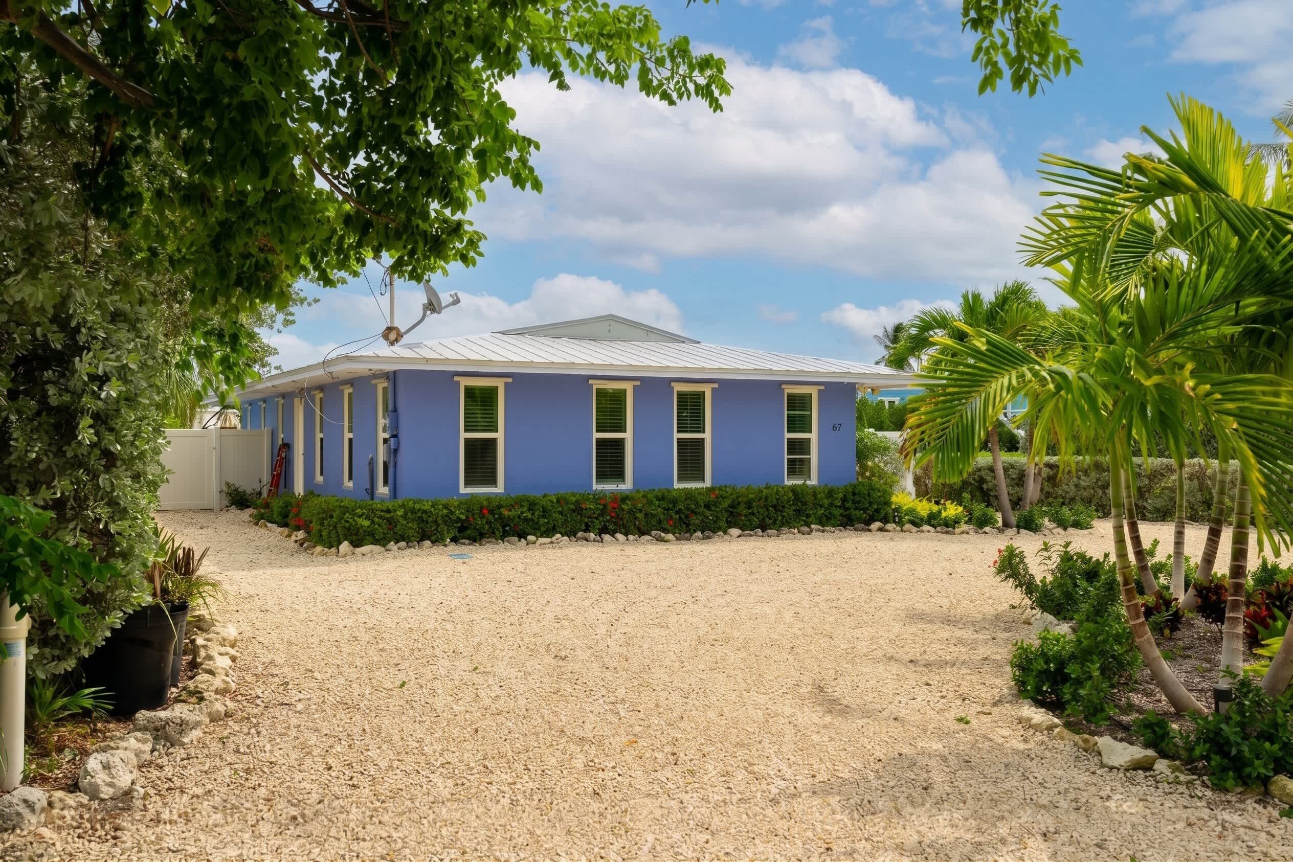 A two-story building with a blue exterior, a gravel driveway, and a well-maintained garden with various plants and a palm tree.